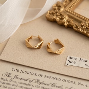 May include: A pair of gold-toned hoop earrings featuring a geometric, angular design with a polished finish. The earrings are shaped like irregular pentagons with a hinged closure mechanism, showcasing a metallic lustre and a structured form.