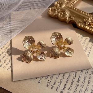 May include: A pair of gold-colored flower-shaped stud earrings. Each earring features four textured petals and a small, round pearl in the center. The earrings are displayed on a beige surface with a vintage aesthetic.