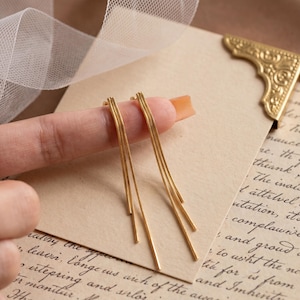 Könnte beinhalten: Goldfarbene Hängeohrringe mit linearem Design. Die Ohrringe bestehen aus mehreren dünnen, geraden Strängen, die herabhängen. Die Ohrringe werden auf einem Finger vor dem Hintergrund eines Vintage-Papiers mit handschriftlichem Text und einer dekorativen Ecke präsentiert.