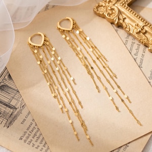 Pode incluir: Um par de brincos pendentes em tom dourado com um topo em forma de gota e várias correntes longas e delicadas penduradas. Cada corrente é adornada com pequenos discos metálicos dourados, planos e refletores, espaçados uniformemente.