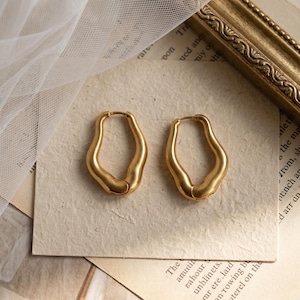 May include: A pair of gold-toned hoop earrings featuring an irregular, organic, and fluid shape with a smooth matte finish. The earrings are designed as continuous loops with a hinged closure mechanism, showcasing a soft metallic luster.