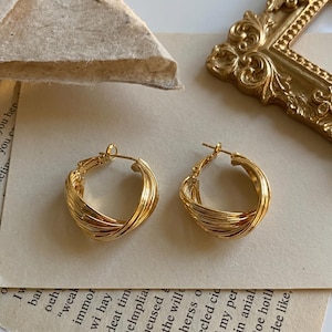 May include: A pair of gold-toned hoop earrings featuring a multi-layered, twisted design. Each earring is constructed from several thin, polished metal strands that overlap to create a continuous, rounded shape with a secure hinged post closure.