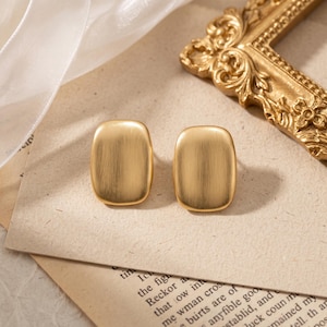 May include: A pair of gold-toned stud earrings featuring a smooth, brushed metal finish and a rounded rectangular shape. The earrings have a soft, matte texture and a simple, clean design that highlights the metallic surface and curved edges.