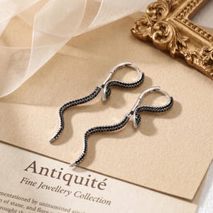 May include: A pair of silver hoop earrings shaped like snakes, featuring black pavé stones along the body and small green stones for the eyes. The earrings have a hinged closure mechanism and are displayed on a card labeled Antiquité.