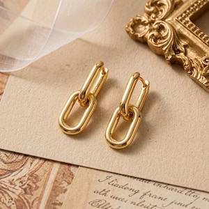 May include: A pair of gold-toned drop earrings featuring a double-link chain design. Each earring consists of two interlocking oval-shaped loops with a polished, smooth finish. The metal surface reflects light, and the hallmark S925 is visible.