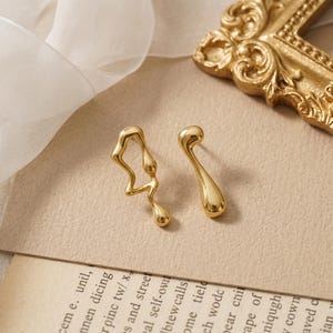 May include: A pair of gold-toned earrings featuring an asymmetrical design. One earring has an open, irregular, fluid shape, while the other is a solid, smooth, teardrop-like form. Both pieces have a polished, reflective metallic finish.