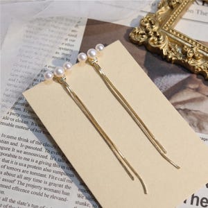Peut inclure: Boucles d'oreilles dorées avec trois fausses perles et de longues chaînes pendantes. Elles sont présentées sur une carte de couleur neutre, mettant en valeur leur design et leur élégance.