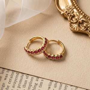 May include: A pair of gold hoop earrings featuring a row of small, round, vibrant red stones set along the outer edge. The earrings are crafted with a polished gold finish and include a 925 hallmark stamp, indicating sterling silver base metal.