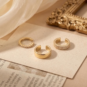 May include: A set of three gold-toned ear cuffs. Two of the cuffs feature a single row of small, clear, sparkling stones embedded along the outer surface, while the central cuff is a plain, smooth, polished gold-toned band with an open design.