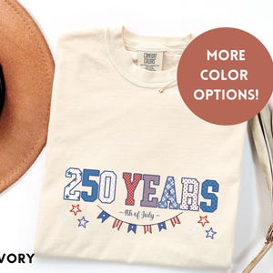 Puede incluir: Camiseta de color marfil con el texto "250 YEARS - 4th of July" en rojo, blanco y azul. El diseño incluye estrellas y una pancarta. Un sombrero marrón es visible en la esquina superior izquierda, y una planta seca está en la esquina derecha.