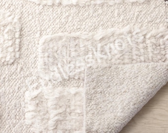 Könnte beinhalten: Ein cremefarbener Teppich mit einem strukturierten, getufteten geometrischen Muster aus abwechselnden Hochflor- und Niederflorabschnitten. Das Wort "lessknots" ist in einer hellen, halbtransparenten Schrift auf der Teppichoberfläche gedruckt.