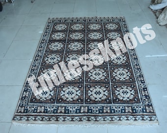 Könnte beinhalten: Ein rechteckiger Teppich mit einem Gittermuster aus dunkelbraunen und cremefarbenen Quadraten. Jedes Quadrat enthält ein zentrales kreisförmiges Blumenmotiv. Der Teppich hat eine dekorative Bordüre mit geometrischen Mustern und kurze Fransen.