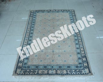 Könnte beinhalten: Ein rechteckiger Teppich mit einem beigen Feld und einem sich wiederholenden Muster aus kleinen blauen geometrischen Motiven. Der Teppich ist von einer breiten Bordüre in Blau- und Beigetönen mit komplexen Mustern umrahmt und enthält den Text EndlessKnots.