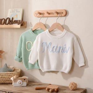 Può includere: Due maglioni in maglia a maniche lunghe appesi a grucce di legno. Il maglione davanti è bianco con la parola "Mani" ricamata in corsivo azzurro. Il maglione dietro è verde chiaro con un dettaglio ricamato circolare. Entrambi hanno colletti a coste.