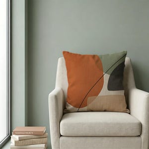 May include: A square throw pillow featuring an abstract design with a large burnt orange circle, a sage green shape, a dark charcoal gray form, and a tan rectangle, all accented by a thin black diagonal line on a light beige textured fabric.