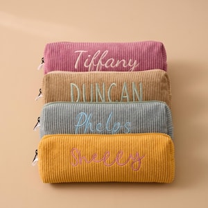 May include: A stack of four rectangular corduroy pencil cases in pink, tan, grey, and mustard yellow. Each case features a side zip and is embroidered with a name in a contrasting thread colour: Tiffany, DUNCAN, Phelps, and Shelly.