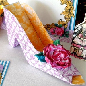 May include: A purple and white paper shoe with a yellow interior and a pink rose on the side. The shoe is a party favor or decoration.