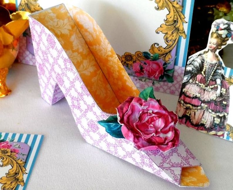May include: A purple and white paper shoe with a yellow interior and a pink rose on the side. The shoe is a party favor or decoration.