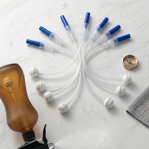 May include: An amber-colored bottle with a black spray nozzle and several clear tubes with blue tips and white bulbous ends. A small brass compass and a gray cloth are also present. The items are arranged on a white marble surface.