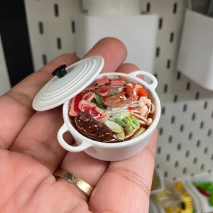Op de afbeelding: Een miniatuurpan in witte keramiekstijl met twee handvatten en een bijpassend deksel, gevuld met een gedetailleerde voedselsculptuur. Het gerecht bevat diverse ingrediënten zoals gesneden paddenstoelen, groene groenten en vlees in een donkere bouillon.
