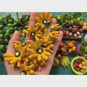 May include: A hand displaying several clusters of miniature yellow bananas. The bananas are arranged in a flower-like pattern. Other miniature fruits and vegetables, including green tomatoes, are visible in the background.