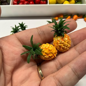 Könnte beinhalten: Zwei Miniatur-Ananas-Figuren mit texturierten gelben und orangefarbenen Fruchtkörpern, die mit spitzen grünen Kunststoffblättern versehen sind. Diese kleinen Dekorationsartikel sind detailreich gestaltet und weisen eine unebene Oberfläche sowie leuchtende Farben auf.