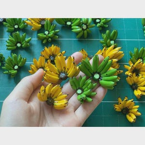 Könnte beinhalten: Miniatur-Bananen in verschiedenen Reifegraden, von grün bis gelb. Die kleinen Bananen sind in Büscheln angeordnet, einige werden in einer Hand gehalten. Die detaillierten, realistischen Bananen liegen auf einer grünen Oberfläche, was auf ein Bastel- oder Lebensmittelthema hindeutet.