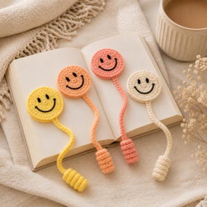 Puede incluir: Cuatro marcapáginas de ganchillo con diseños circulares de caras sonrientes en amarillo, naranja, coral y crema. Cada marcapáginas tiene un tallo largo, fino y flexible que termina en un peso cilíndrico acanalado sobre un libro abierto.