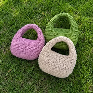 May include: Three small, rounded handbags crafted from crocheted yarn. One bag is light purple, one is bright green, and one is a neutral beige color. Each bag features a single integrated handle and a dense, textured stitch pattern throughout.