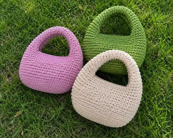May include: Three small, rounded handbags crafted from crocheted yarn. One bag is light purple, one is bright green, and one is a neutral beige color. Each bag features a single integrated handle and a dense, textured stitch pattern throughout.