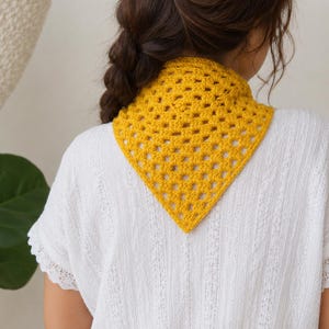 Può includere: Una bandana triangolare all'uncinetto di colore giallo brillante con un classico motivo a piastrelle granny. L'accessorio è indossato intorno al collo e presenta un design traforato e testurizzato con un bordo inferiore a punta e un motivo ripetuto.