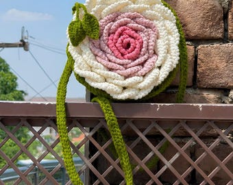 May include: A round crochet bag shaped like a large flower with layered white and pink petals. The bag features a green crochet strap and two small green leaves attached to the side. The design mimics a blooming rose with a pink center.
