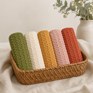 May include: A rectangular woven basket containing five rolled crochet washcloths in olive green, cream, mustard yellow, dusty pink, and terracotta orange. Each washcloth features a consistent textured stitch pattern arranged side by side.