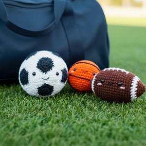 May include: Crocheted sports balls including a soccer ball, basketball, and football, each with smiling faces. The soccer ball is white with black patches, the basketball is orange, and the football is brown with white stripes. A navy blue duffel bag is in the background.