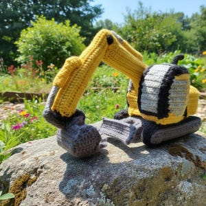 Puede incluir: Un juguete de excavadora hecho a mano con ganchillo en amarillo, gris y negro. El juguete está sobre una gran roca con un fondo verde y floral. La excavadora presenta un diseño detallado con un brazo y un cucharón móviles.