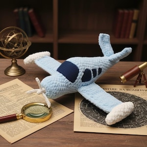 Puede incluir: Un avión de juguete de ganchillo azul claro con detalles blancos en las puntas de las alas y la hélice. El avión presenta una cabina azul oscuro y se exhibe sobre una superficie de madera con accesorios vintage.