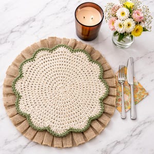 Puede incluir: Un mantel individual de ganchillo color crema con un borde festoneado, bordeado por costuras verdes y un volante de arpillera. Una vela encendida en un tarro de cristal marrón, un pequeño jarrón de flores, un tenedor, un cuchillo y una servilleta estampada completan la mesa.
