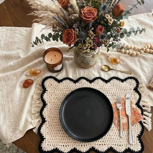 Może przedstawiać: Nakrycie stołu z czarnym talerzem, sztućcami i kwiatową serwetką na kremowej, szydełkowej podkładce z falistymi brzegami. Na stole znajduje się również wazon z kwiatami, świeca i kryształy.