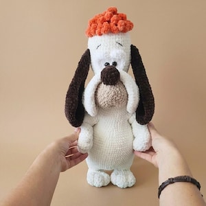 May include: A handmade, plush dog toy in white, brown, and orange. The dog has long, floppy brown ears, a brown nose, and an orange, textured hat. The toy is held by two hands.