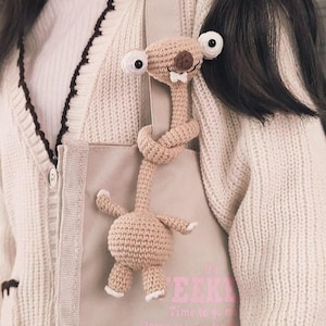 May include: A beige crocheted sloth toy with large white eyes and a small white bow tie. The sloth is attached to a beige tote bag. The tote bag has text that reads "EEK! Time to go make some stories for..."