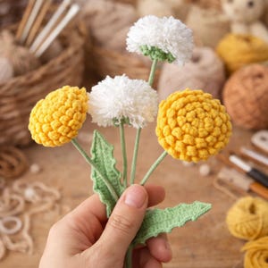 May include: A hand holds a bouquet of crocheted flowers. The bouquet features three yellow and two white fluffy flower heads with green stems and leaves. The flowers are handmade and appear to be crafted from yarn.