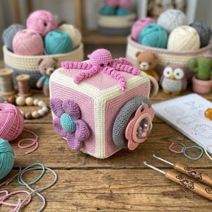 Può includere: Un cubo giocattolo all'uncinetto fatto a mano in rosa e bianco con un polpo rosa sopra. Il cubo presenta un fiore e un elemento simile a un ciuccio. Altri oggetti all'uncinetto e gomitoli di lana sono sullo sfondo.