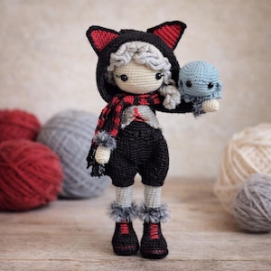 Puede incluir: Muñeca de ganchillo hecha a mano que lleva una chaqueta con capucha negra con detalles rojos y orejas de gato, una bufanda roja y negra y pantalones cortos negros. La muñeca sostiene un pequeño pulpo azul. Las botas de la muñeca son negras con cordones rojos.