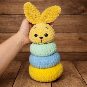 Puede incluir: Un juguete de conejo de ganchillo amarillo, azul y verde. El conejo tiene orejas grandes y un diseño apilable. Está hecho de hilo suave y texturizado, con ojos de seguridad negros y una nariz y boca cosidas.