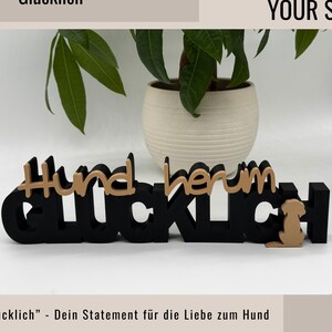 Ein Aufsteller mit den Worten "Hundherum Glücklich" in Schwarz und Hellbraun, mit einer Hunde-Silhouette. Das Schild wird vor einer Topfpflanze mit grünen Blättern angezeigt. Der Text oben lautet "WE INSPIRE YOUR STYLE!" Hundherum glücklich  - Dein Statement für die Liebe zum Hund.