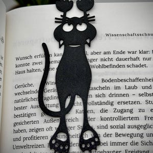 May include: A black cat bookmark, with a smiling face and flower-like shapes on its head, is placed in a book. The bookmark's design includes whiskers and paws. The book's pages are visible, with German text.