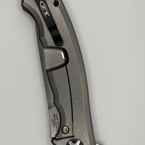 May include: A gray folding knife with a black pocket clip. The knife has a textured handle and a blade. The text "ZT" is visible on the clip. The text "0452CF Sinkevich Design USA S35VN" is also visible.