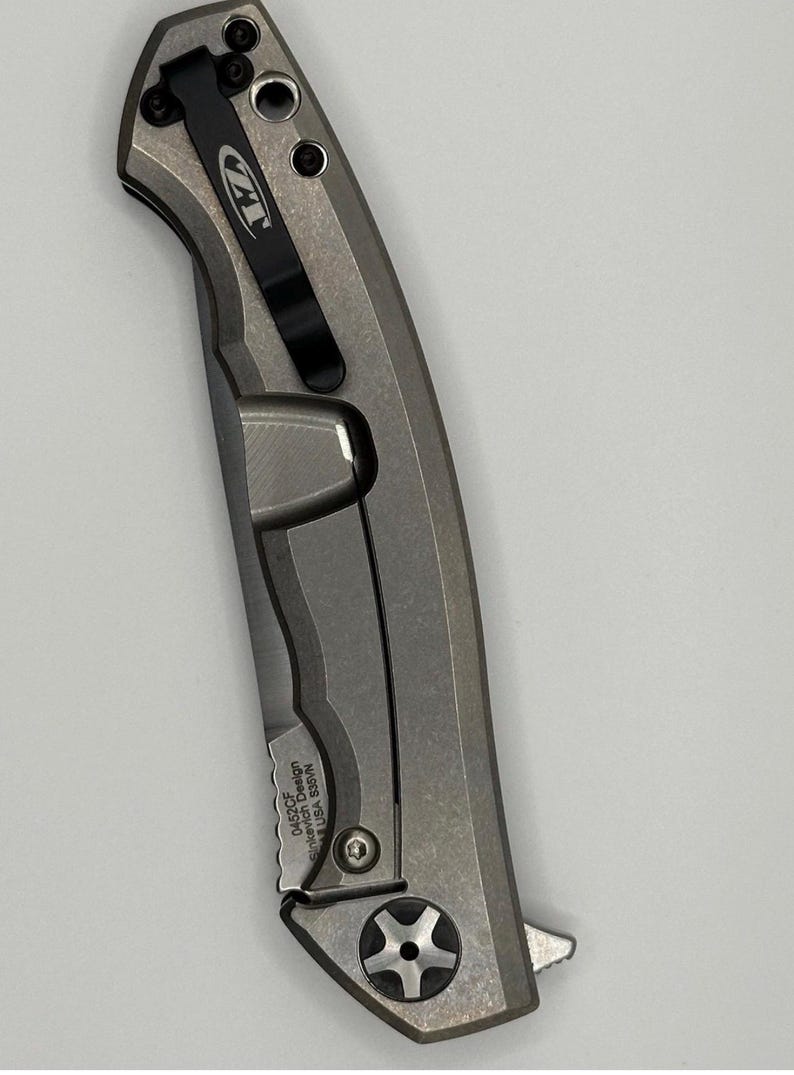 May include: A gray folding knife with a black pocket clip. The knife has a textured handle and a blade. The text "ZT" is visible on the clip. The text "0452CF Sinkevich Design USA S35VN" is also visible.