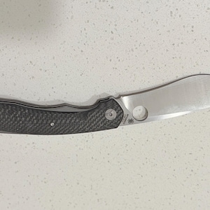 May include: A silver folding knife with a curved blade and a dark grey textured handle. The blade has a circular hole near the handle. The handle is made of a dark grey carbon fibre material.