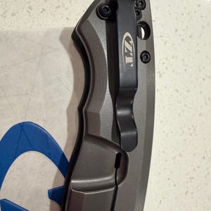 May include: A gray and black folding knife with a pocket clip. The knife has a textured handle and a black clip with the letters "ZT" engraved on it. The blade is not visible in the image.